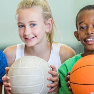 Fem barn håller glada i olika sportbollar, som fotboll, volleyboll och basket, vilket skapar en livlig atmosfär i ett väl upplyst utrymme inomhus.
